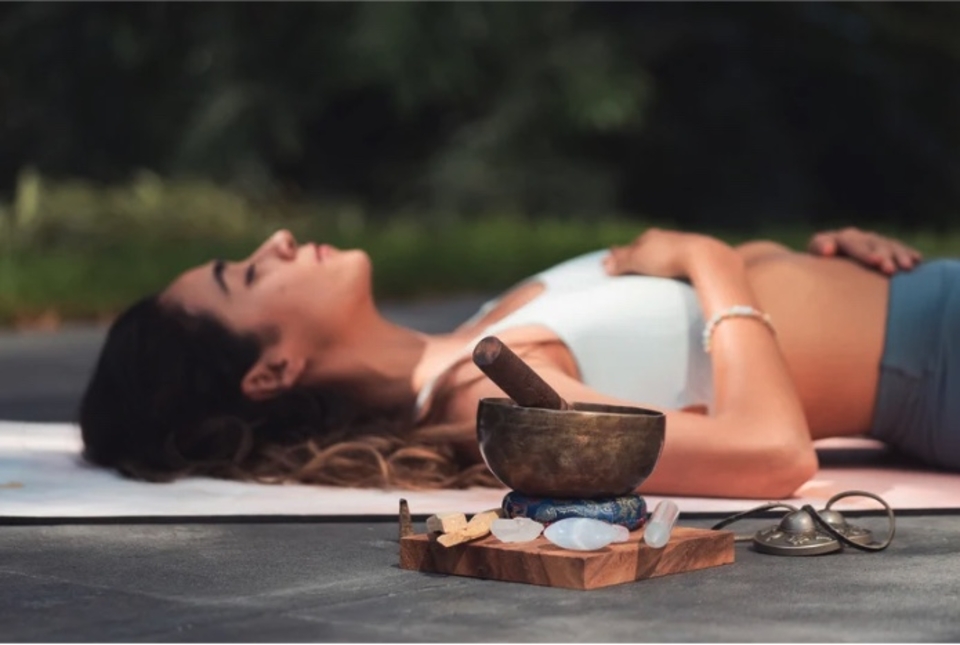 Frau liegt am Rücken im Freien auf einer Yogamatte, eine Hand am Bauch, die andere auf der Brust, neben ihr Klangschale auf einem Holzbrett, Peggy Böhmer